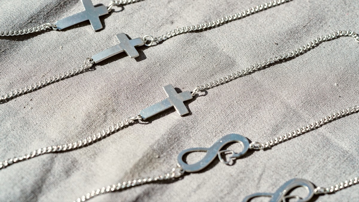 Bracelets-chaînes à pendentifs croix et infini en acier inoxydable couleur argent disposés sur du lin gris